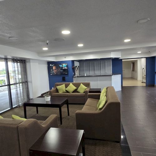 The image shows a spacious lounge area with sofas, a coffee table, and large windows, creating a welcoming and well-lit atmosphere.