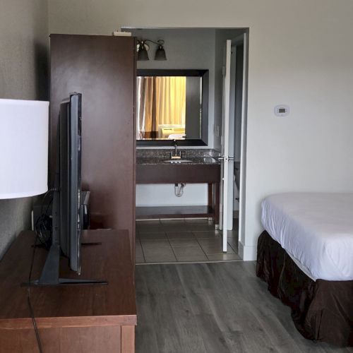 A hotel room with a bed, TV on a dresser, mirror, and bathroom in the background. The room has wooden flooring and a simple design.