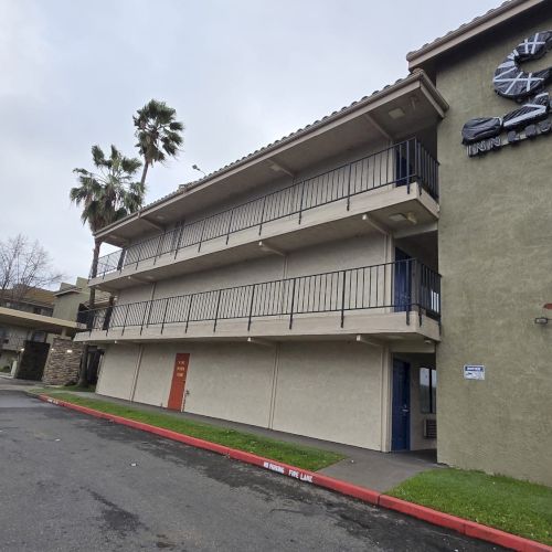 The image shows a multi-story motel building with exterior corridors, a sign on the wall, and palm trees nearby.