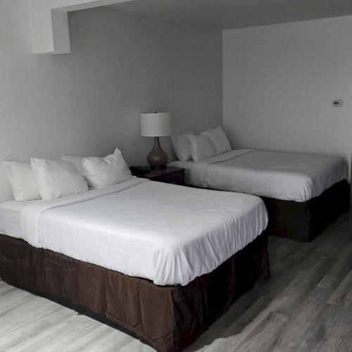 The image shows a hotel room with two neatly made beds, a nightstand with a lamp between them, and light wood flooring.