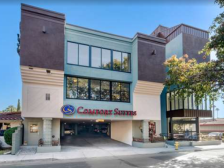 The image shows a building with a "Comfort Suites" sign, featuring a modern design with large windows and trees nearby.