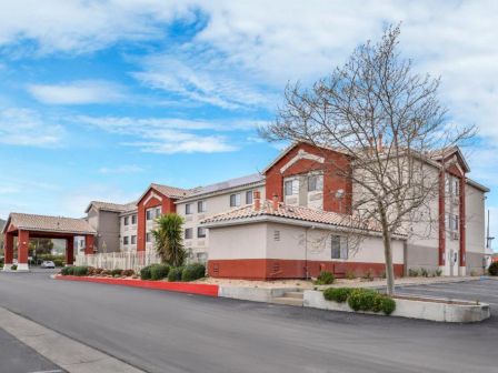 A two-story building with red and beige exterior, a paved driveway, parking spaces, and trees. The sky is partly cloudy.