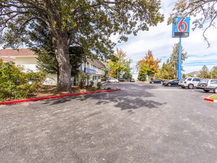 A quiet motel parking lot with trees lining the sides, a few cars parked, and a blue and red sign in the distance.