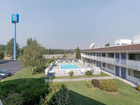 A motel-style courtyard shows a rectangular pool fenced in, a two-story beige building with balconies, a parking lot, trees, and green lawns.