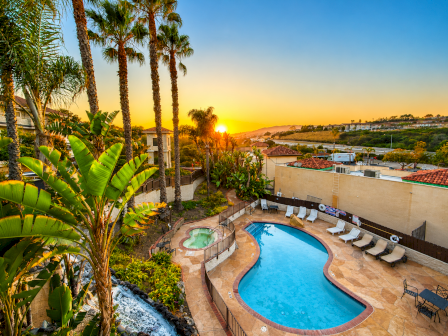 A resort pool area with palm trees, sun loungers, and a curved blue pool at sunset, surrounded by greenery and warm lighting.