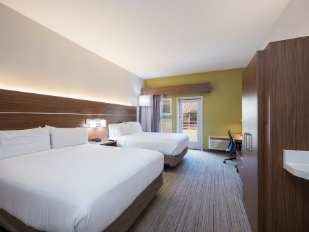 A modern hotel room with two large beds, a desk, a chair, a wardrobe, and a window letting in natural light.