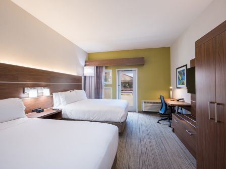 A hotel room with two queen beds, a wooden headboard, nightstands, a work desk, chair, wardrobe, and a door leading to a balcony, all in a bright, modern layout.