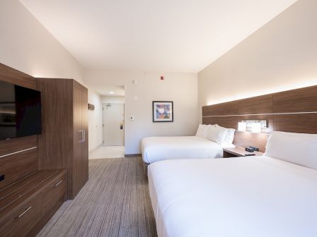 Two double beds in a modern hotel room with a wooden dresser, wall art, and a TV; neutral tones and soft lighting create a calm vibe.