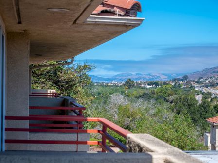 A balcony view from a hillside apartment shows a concrete railing, partial building edge, lush green trees, distant hills, and a blue sky over a valley.