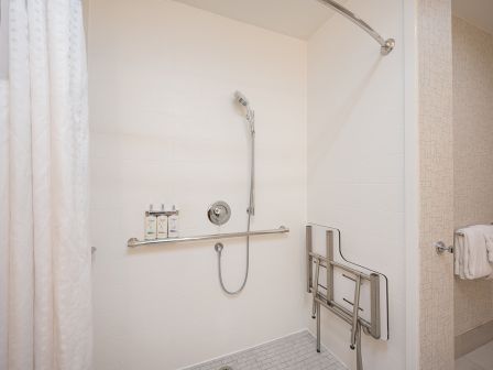 A modern accessible shower with a handheld shower, a fold-down grab bar, a shower chair/rail and a textured floor, with a curtain on the left side.