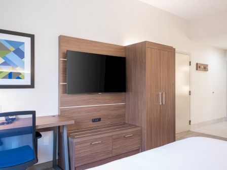A modern hotel room with a wall-mounted TV, wooden cabinet and desk, blue chair, abstract framed art, and a bright doorway to the hallway.