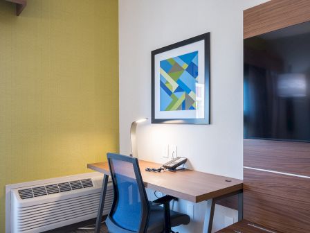 A compact hotel desk nook with a wooden desk, blue office chair, framed geometric art, a wall lamp, and a green accent wall beside an air conditioner.