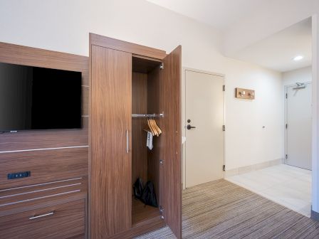A modern hotel room with a wall-mounted TV, an open wooden wardrobe showing hangers, a door, and a light-colored hallway leading to the entrance.
