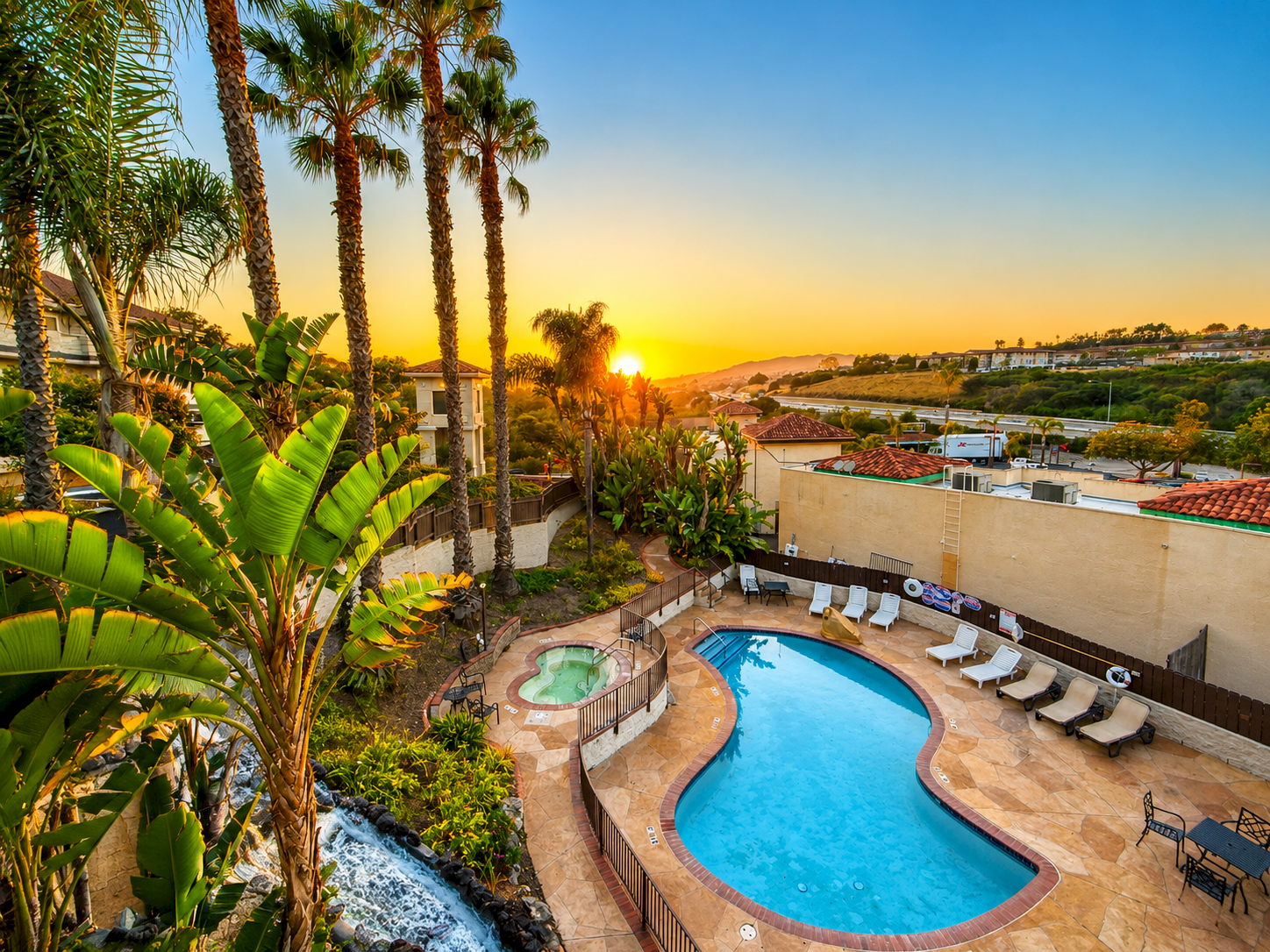 A sunny poolside view with a curvy blue pool, lounge chairs, tall palm trees, and a vibrant sunset in the background.