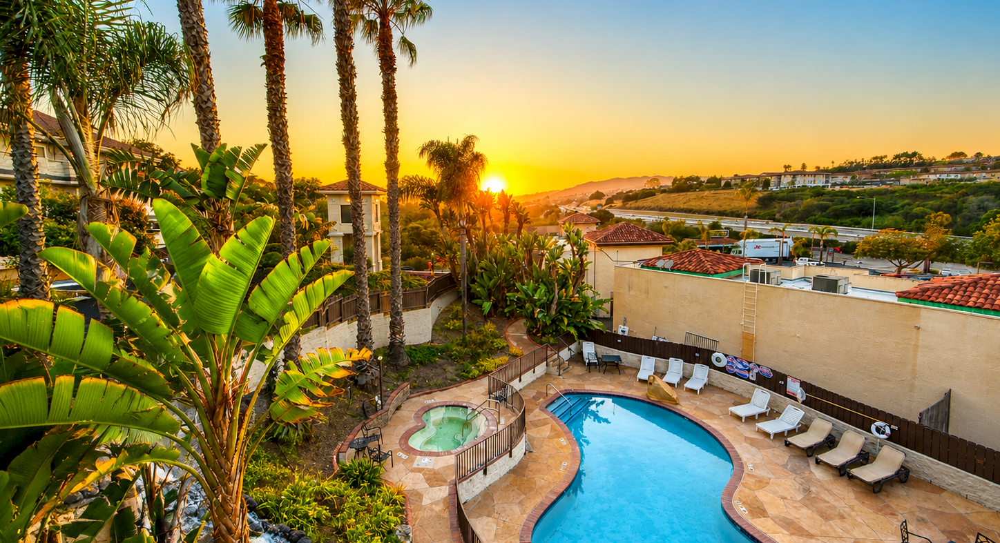 A sunny poolside view with a curvy blue pool, lounge chairs, tall palm trees, and a vibrant sunset in the background.