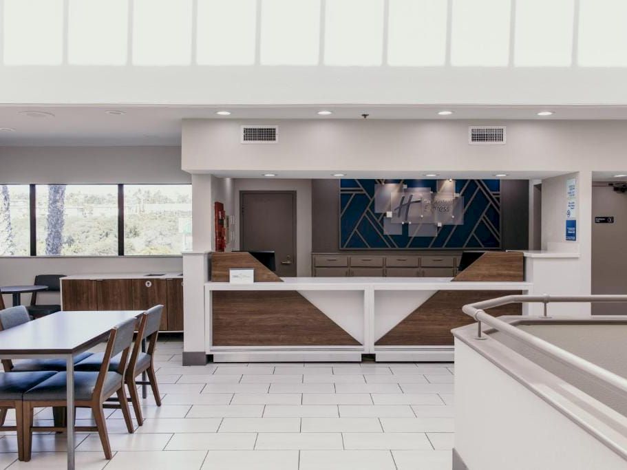 A bright hotel lobby with a check-in desk, seating area, white tile floor, and modern geometric wall decor in the background.