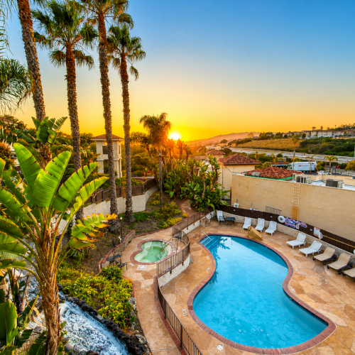 Luxurious resort pool with curvy blue water, sun loungers, lush palm trees, and a golden sunset over a terraced hillside retreat.