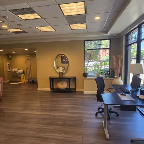 A bright office waiting area with chairs, a reception table, large windows, and a mirrored wall, set in a quiet hallway.