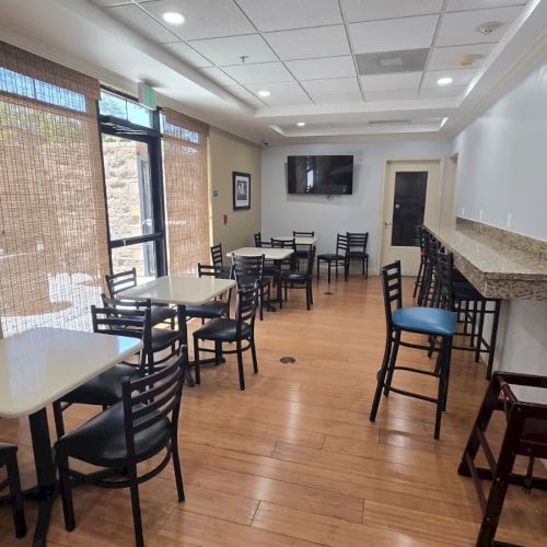 A bright break room/caf&eacute; with several tables and chairs, big windows with blinds, a wall TV, and a granite counter along the side.