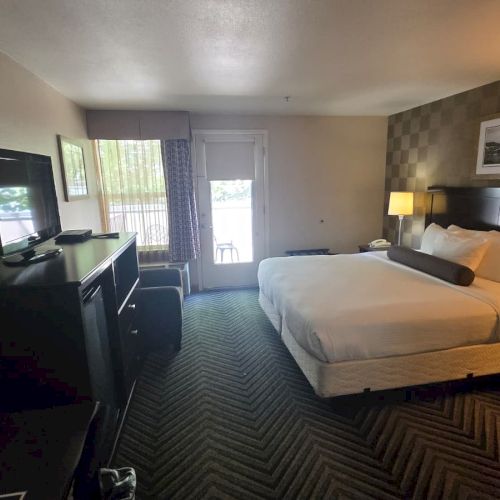 A hotel room with a neatly made bed, bedside lamps, a desk with TV, and curtains and windows letting in daylight.