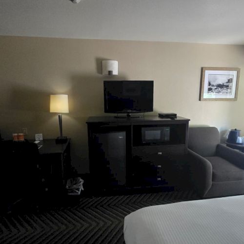 A hotel room with a TV on a cabinet, sofa, desk and lamps, plus a window with curtains and a small kettle.