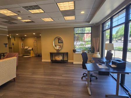 An office reception area with chairs, a desk, framed art, large windows, and a round wall mirror in a bright hallway,
