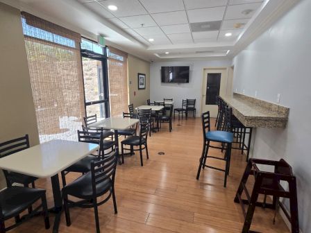 A small indoor dining room with tables, black chairs, a granite bar counter, and a wall-mounted TV, viewed through blinds.