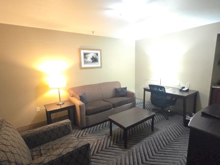 A cozy hotel room with a sofa, armchairs, coffee table, lamp, desk, filing cabinet, and a TV, all neatly arranged.