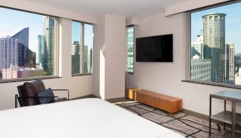 A modern hotel room with large windows showcases a cityscape. Features include a bed, armchair, wall-mounted TV, and sleek furniture.