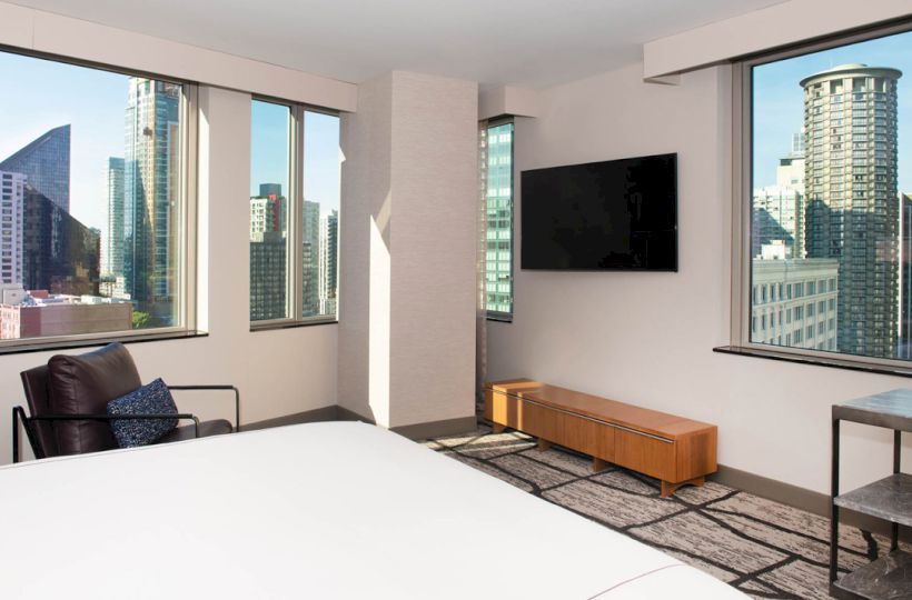 A modern hotel room with large windows showcases a cityscape. Features include a bed, armchair, wall-mounted TV, and sleek furniture.