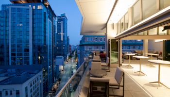 Rooftop patio with tables and chairs overlooking a cityscape of modern high-rise buildings at dusk, warmly lit interior visible inside.