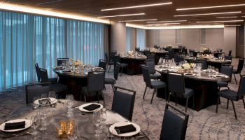 The image shows an elegantly set banquet room with round tables, chairs, glassware, and napkins, illuminated by modern ceiling lights.
