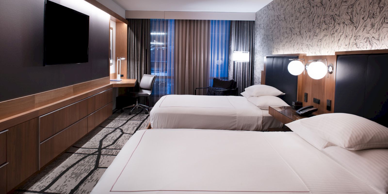 The image shows a hotel room with two neatly made beds, a wall-mounted TV, a desk, a chair, and modern decor including patterned wallpaper.