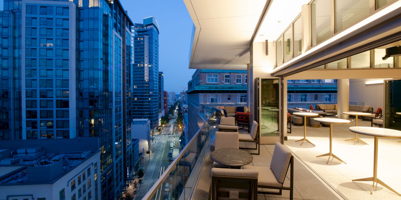 A modern balcony overlooking a city street at dusk, with tables and chairs. Buildings with illuminated windows line the street.