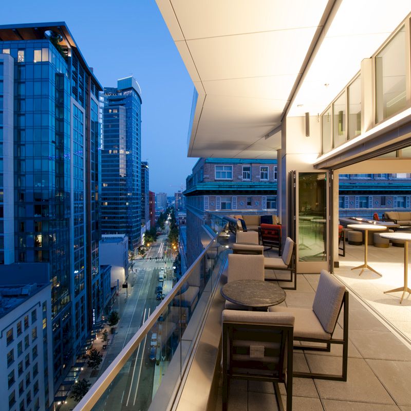 A modern balcony overlooking a cityscape at dusk, with tables and chairs. Skyscrapers and streets are visible under the evening sky.