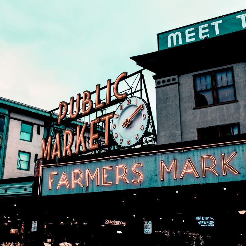 The image shows a neon sign for a "Public Market" and "Farmers Market" on a building, featuring a clock.