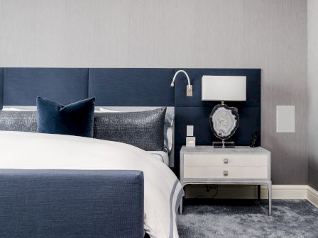 A modern bedroom with a blue headboard, white bedding, side table, lamp, and decorative art piece; floor covered with a plush gray carpet.