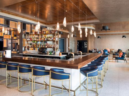A modern bar with a central counter, bar stools, hanging lights, and shelves stocked with bottles. Seating areas are visible in the background.