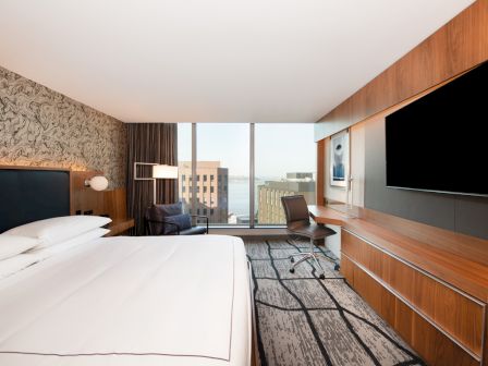 A modern hotel room with a large bed, work desk, and expansive window view of city buildings and water, featuring stylish decor and a TV.