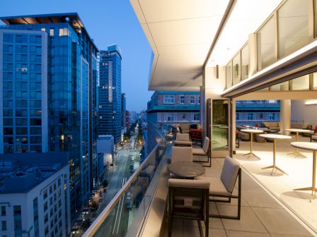 A modern rooftop terrace with tables and chairs overlooks a cityscape at dusk, featuring tall buildings and a clear sky.