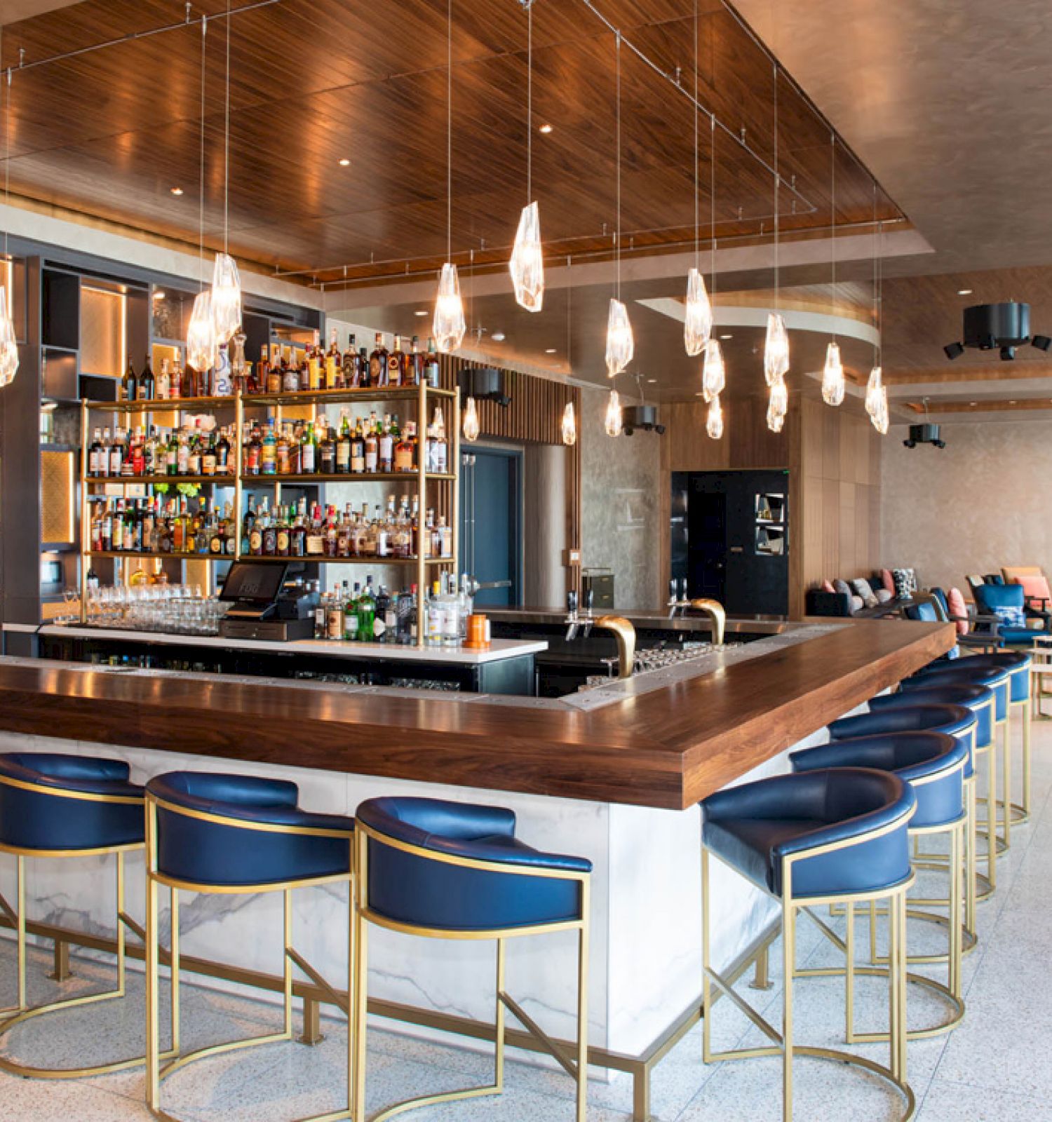 A modern bar with blue stools, pendant lights, and a well-stocked liquor shelf. Cozy seating and a neutral color scheme enhance the ambiance.