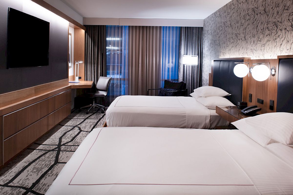A modern hotel room with two beds, a large TV, a desk, and stylish lighting. The decor is contemporary with patterned wallpaper and carpet.