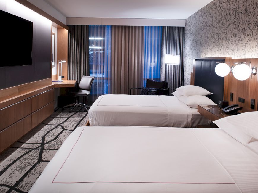 A modern hotel room with two beds, a large TV, a desk, and stylish lighting. The decor is contemporary with patterned wallpaper and carpet.