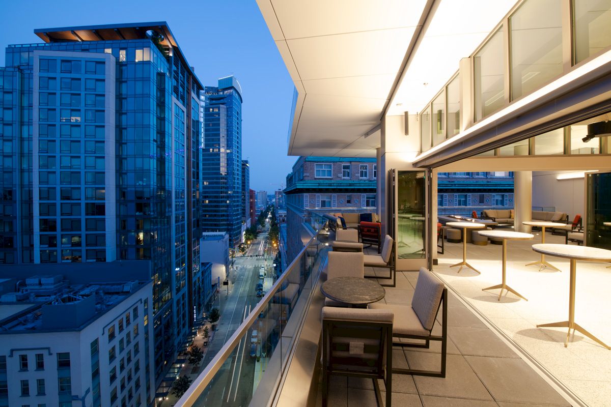 A modern city view from a stylish rooftop patio, featuring tables, chairs, and ambient lighting, all overlooking the bustling streets below.