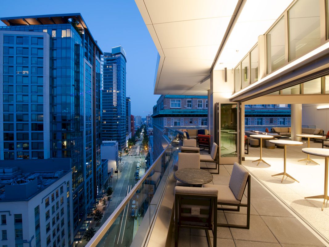A modern city view from a stylish rooftop patio, featuring tables, chairs, and ambient lighting, all overlooking the bustling streets below.