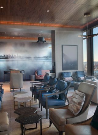 The image shows a modern lounge with plush chairs, a city skyline mural, and large windows offering a view of water and a Ferris wheel.