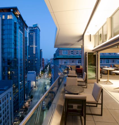 A modern cityscape at dusk with a view from an outdoor terrace featuring tables and chairs overlooking a street with tall buildings.