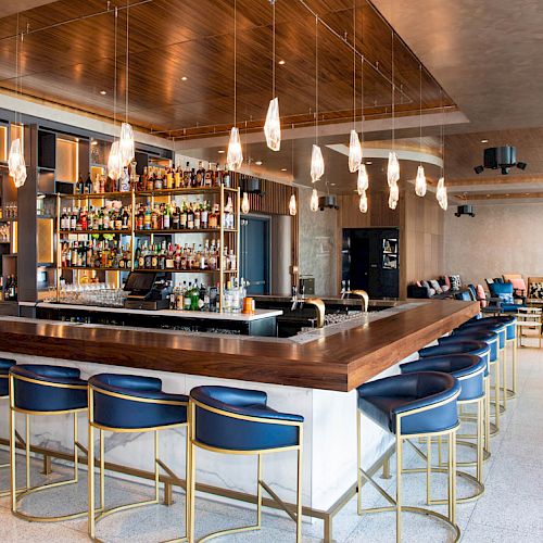 A chic, modern bar features a counter with blue chairs, hanging lights, and a well-stocked shelf. Cozy seating is arranged in the background.