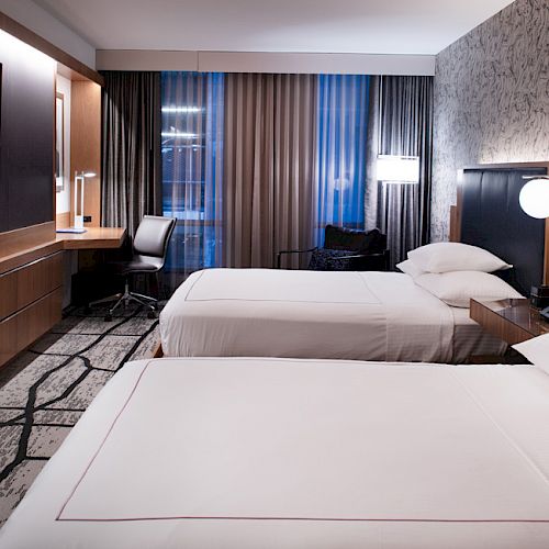 The image shows a modern hotel room with two beds, a TV, a desk, and lamps beside the beds on a patterned carpet.