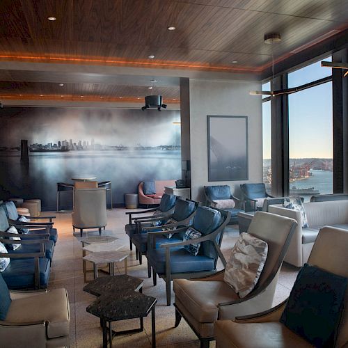A cozy lounge area with modern seating, large windows offering a view of water and a Ferris wheel, and a cityscape mural on the wall.